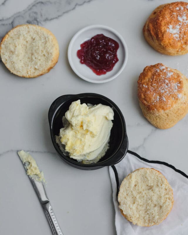 An overhead view of Cornish clotted cream teas with freshly baked scones, some halved and ready to be topped, accompanied by a dish of cornish clotted cream, a small plate of red strawberry jam, and a butter knife. This simple, elegant flat lay captures the essence of national cream tea day, celebrating the beloved British tradition in style.