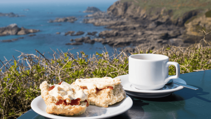A plate of scones topped with jam and clotted cream sits beside a cup of tea on a table overlooking a stunning coastal view of rocky cliffs and blue sea, capturing the perfect British seaside moment. This idyllic scene celebrates national cream tea day with a classic cream tea setup in a scenic outdoor setting.