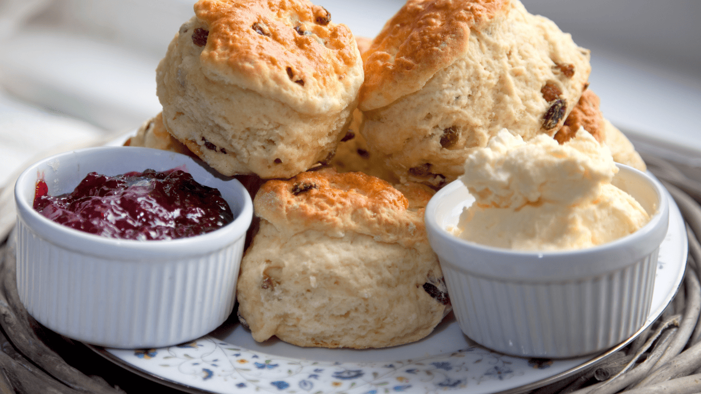 A close-up of a plate filled with golden, fruit-studded scones, served alongside ramekins of rich clotted cream and glossy strawberry jam. This inviting setup is a classic tribute to national cream tea day, showcasing the essential elements of a traditional cream tea.